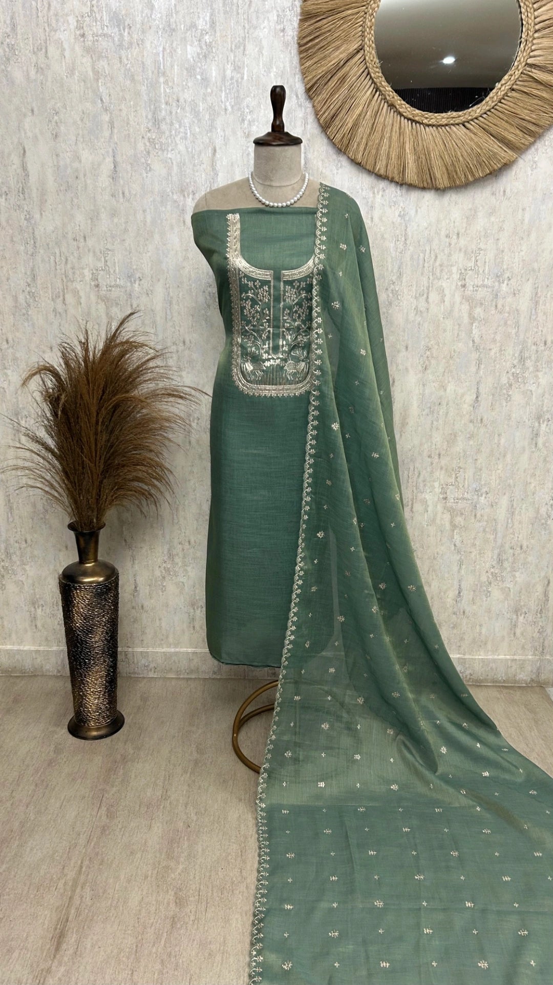 Green embroidered dress on a mannequin with a decorative mirror and vase in the background.